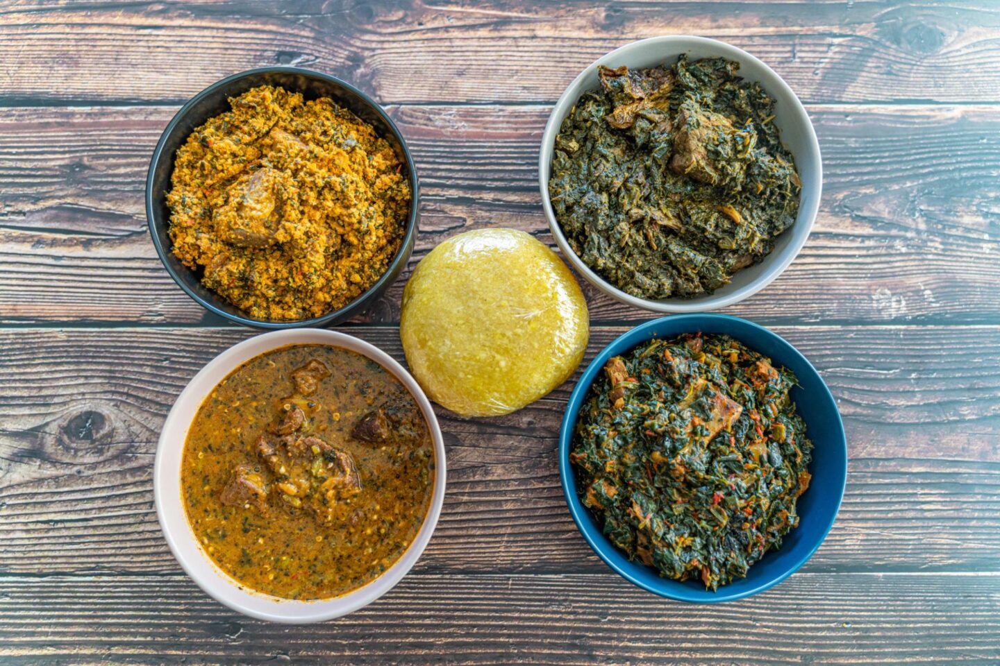 A top-down view of traditional Nigerian dishes including eba and various soups served in bowls on a wooden table.