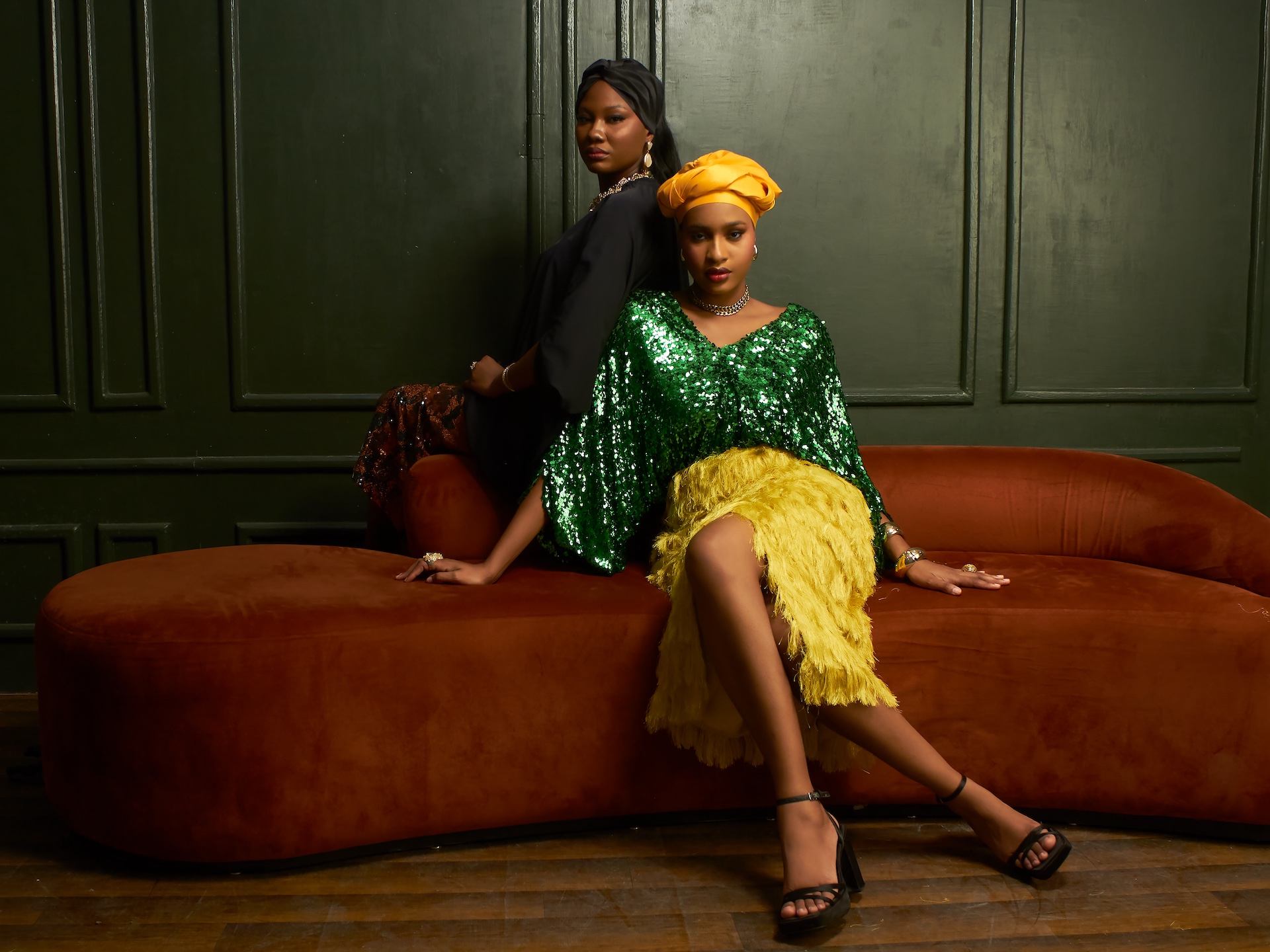 Two women posing stylishly in contemporary African fashion against a dark green paneled wall.