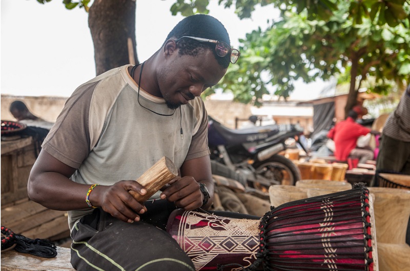 A craftsman carefully carves and assembles a traditional drum in an outdoor workshop.