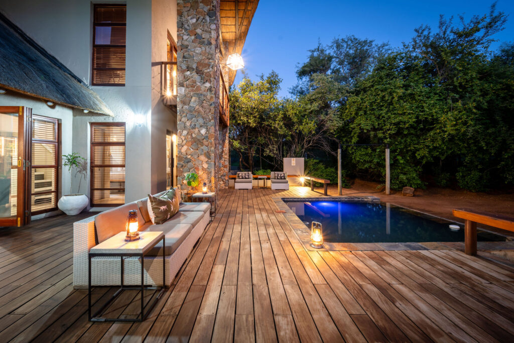 An exterior view of a luxury bush lodge wooden deck with a swimming pool and seating area at dusk in Kruger National Park, South Africa.