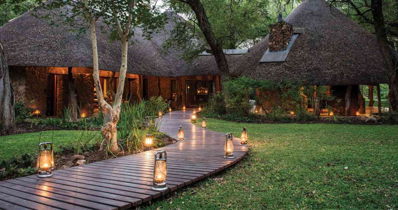 A beautifully illuminated wooden walkway leading to thatched-roof buildings at a luxury safari lodge in South Africa.