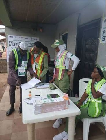 Election officials handling paper documents and ballot boxes inside a room.