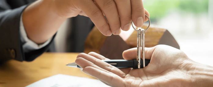 Close-up of hands exchanging house keys in front of a residential home.