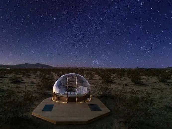 Transparent stargazing glamping dome set on a wooden platform in a desert landscape under a starry night sky.